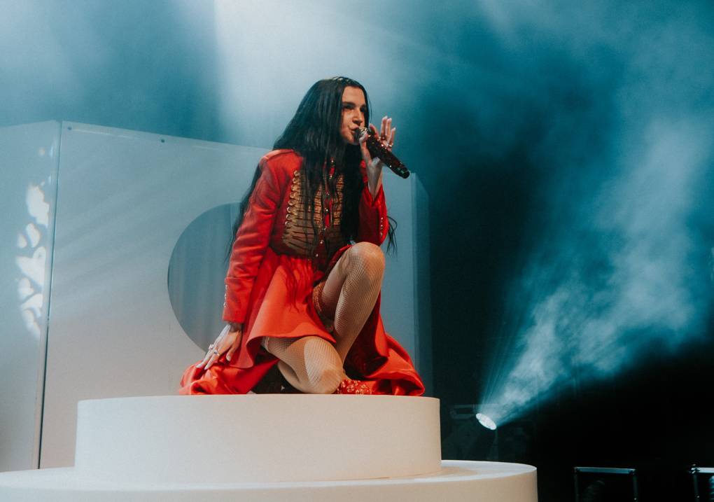 A young woman with long brown hair in a red military-style coat crouches on a white riser, singing into a microphone